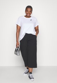 T-shirt blanche à manches courtes avec logo Nike noir, pantalon large noir, sac à main rayé noir et blanc, et baskets montantes noires.