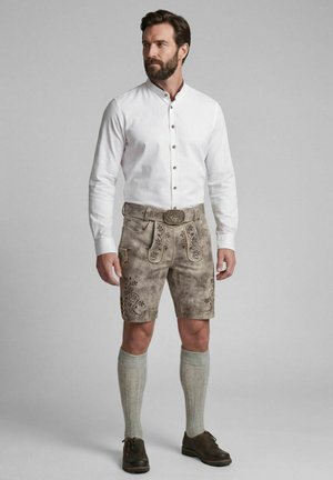 Mann trägt traditionelle beige Lederhose mit Stickerei, ein weißes Hemd mit Knöpfen, kniehohe gestrickte Socken und dunkelbraune Schuhe.