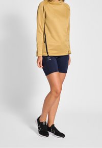 Långärmad huvtröja i beige, tillsammans med marinblå träningsshorts. Svarta detaljer på sidorna och logotypdetalj på shortsen. Svarta skor med vita kanter.