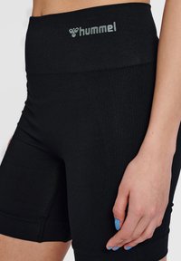 Närbild av en person iklädd svarta Hummel höga träningsshorts, hand vila vid låret med blå målade naglar.