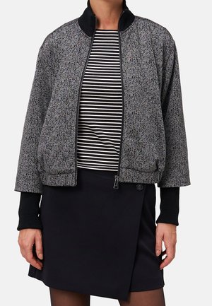 Frau trägt eine schwarz-weiß gemusterte Reißverschlussjacke über einem schwarz-weiß gestreiften Shirt und einem schwarzen Rock mit Knopfdetail.