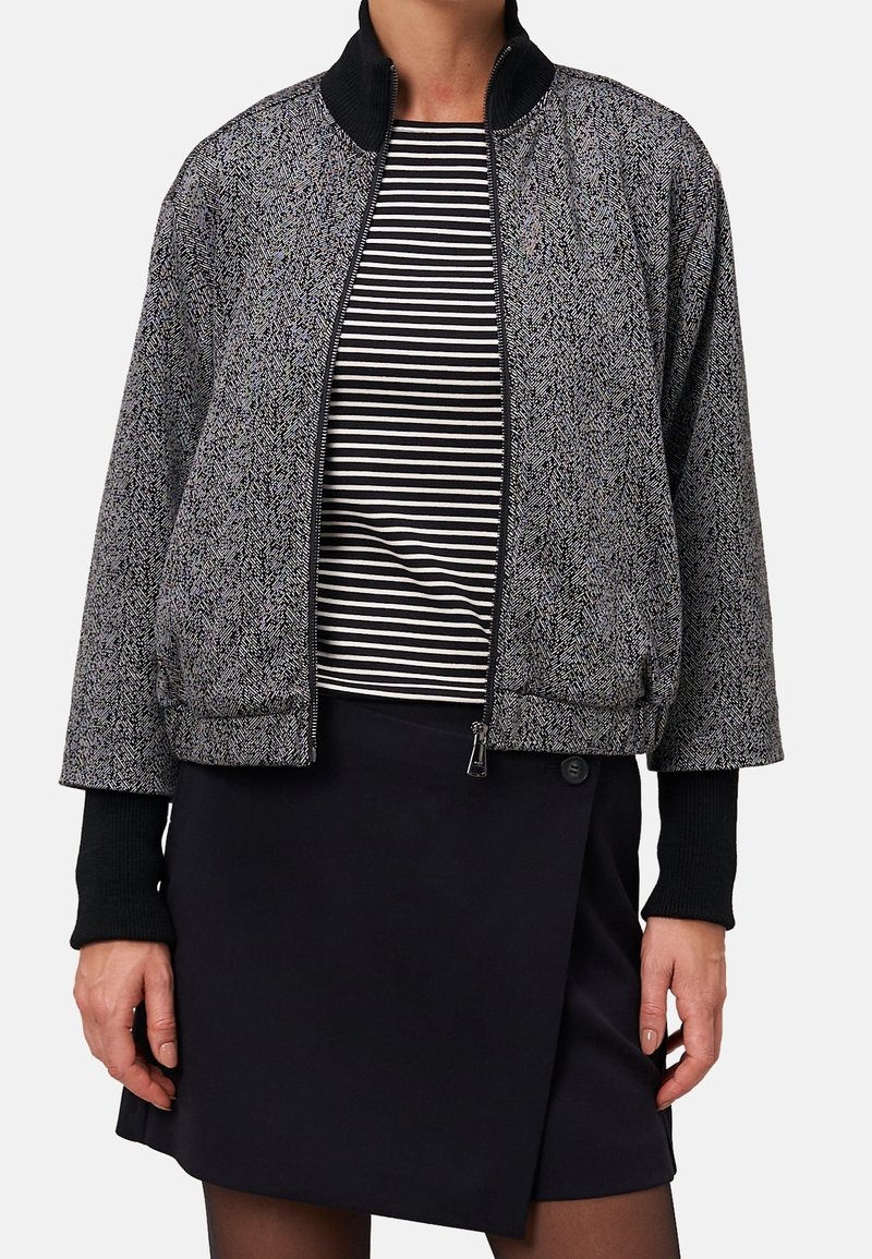 Femme portant une veste zippée à motif noir et blanc sur une chemise rayée noir et blanc et une jupe noire avec un détail bouton.