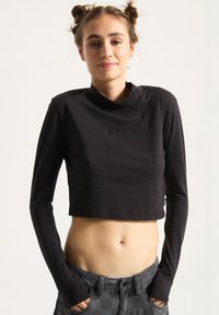 Black cropped long-sleeve top with a high collar, soft fabric, and embroidered logo on the chest. Paired with textured gray checkered pants.