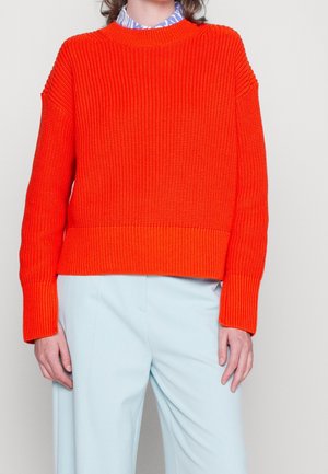 Chunky knitted orange sweater with ribbed texture and long sleeves, featuring a round neckline and relaxed fit, paired with light blue trousers.