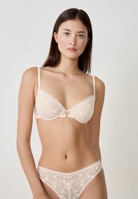 Reggiseno in pizzo e perizoma coordinato in rosa chiaro. Il reggiseno presenta coppe imbottite, design floreali e un fiocco centrale. Tessuto morbido e testurizzato in tutto.