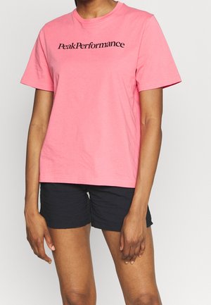 Person wearing a pink short-sleeve shirt with "Peak Performance" text and black shorts, standing against a plain light background.