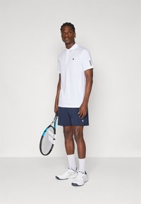 Camisa polo branca com colarinho e logótipo, combinada com calções azul-marinho. A pessoa segura uma raquete de ténis e usa sapatilhas e meias brancas.