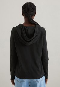Persona con capelli intrecciati raccolti indietro, che indossa una felpa nera con cappuccio e jeans azzurri chiari, girata di spalle su uno sfondo uniforme.