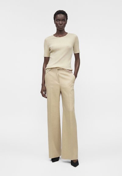 Γυναίκα όρθια με το ένα χέρι στην τσέπη, φοράει μπεζ ριμπ κοντομάνικη μπλούζα, μπεζ παντελόνια με φαρδιά πόδια και μαύρα μυτερά παπούτσια.