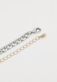 Cadena de metal plateado con un cierre en un extremo, emparejada con una cadena de alambre dorado que presenta eslabones circulares y un pequeño detalle en forma de bola en el otro extremo.