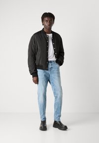 Chaqueta bomber negra con botones a presión, camiseta blanca, jeans ligeros azules y botas negras por el tobillo. El diseño presenta un ajuste holgado y puños acanalados.