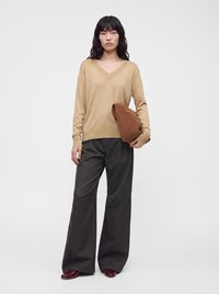 Femme aux longs cheveux noirs portant un pull beige à col en V, un pantalon large noir, des chaussures bordeaux, tenant une pochette pliée marron.