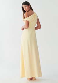 Gul lång klänning med axelbandslös design, flödande tyg och en mjuk textur. Har en markerad midja och en slits i sidan för rörelsefrihet.