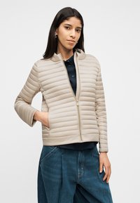 Γυναίκα με σκούρα μαλλιά, ντυμένη με μπεζ καπιτονέ μπουφάν πάνω από σκουρόχρωμο πουκάμισο και φαρδιά μπλε τζιν, στέκεται με το ένα χέρι στην τσέπη του μπουφάν.