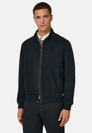 Giovane uomo che indossa una giacca nera con cerniera sopra una camicia bianca con colletto e un maglione marrone chiaro, in piedi contro uno sfondo chiaro e uniforme.