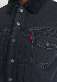 Mörk denimjacka med en fluffig svart krage, kopparknappar och en liten röd etikett på bröstfickan. Strukturerad tyg med synliga sömmar.