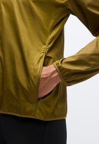 Chaqueta ligera de color verde oliva con diseño aerodinámico, puño elástico y bolsillo lateral en la costura. Textura suave y mate.