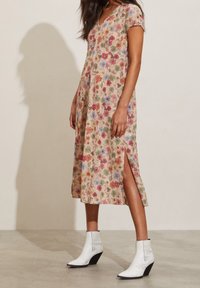 Robe midi fleurie en tissu beige à manches courtes, ornée de motifs floraux multicolores et d'une fente sur le côté. Portée avec des bottes blanches à talons.