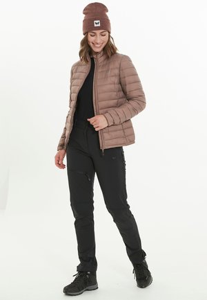 Veste matelassée marron clair avec col montant, associée à un pantalon noir avec poches zippées et des bottes de randonnée noires. Bonnet marron avec logo.