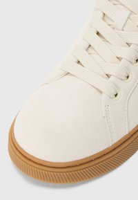 Fehér szarvasbőr sportcipő bézs gumi talppal és lapos fűzőkkel. Lekerekített orr-résszel és finom varrással a fűzőlyukak közelében.