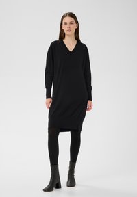 Schwarzes Strickkleid mit V-Ausschnitt, langen Ärmeln und lockerem Sitz. Verfügt über einen gerippten Saum und Seitenschlitze, kombiniert mit schwarzen Stiefeletten.