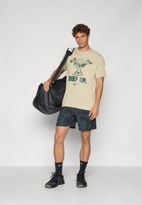 Beige t-shirt med grøn grafik og teksten "BEEF UP," parret med mørke mønstrede shorts. Sort duffel taske og sorte sportssko.