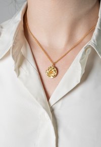 Goldene Halskette mit einem ovalen Anhänger, der florale Muster und kleine grüne Akzente aufweist, an einer Kette, die über einem weißen Hemd mit Kragen getragen wird.