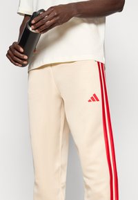 Beige Sporthose mit roten Seitenstreifen und einem Adidas-Logo am linken Oberschenkel; eine Person hält eine schwarze Wasserflasche.
