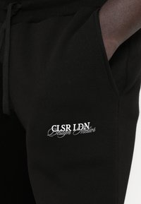 Joggers pretos feitos de tecido macio apresentam cintura com cordão e bolsos laterais, com um logo branco bordado na coxa.