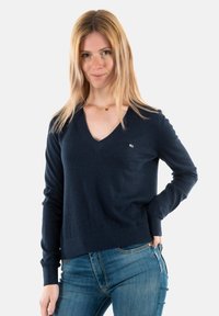 Maglione blu navy a collo a V realizzato in un tessuto morbido, caratterizzato da maniche lunghe e orlo a costine. Include un piccolo dettaglio del logo sul petto.