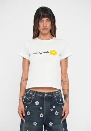 T-shirt branca com texto preto "Marc Jacobs" e um aplique de flor amarela. Usada com calções de ganga escuros featuring embellishments em forma de flor prateada.