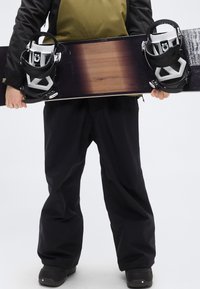 Σνόουμπορντ με σκούρο ξύλινο φινίρισμα και μαύρες δέστρες, κρατιέται όρθιο. Ο αναβάτης φοράει μαύρο παντελόνι και μαύρες μπότες, με πράσινο μπουφάν.