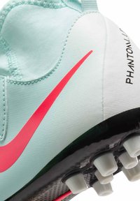 Des chaussures de football bleu clair avec un swoosh rose, une tige en tissu texturé et une semelle noire avec des crampons blancs pour l'adhérence. La marque "Phantom" est visible.