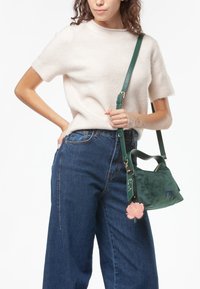Groene suede handtas met metalen hardware, bloemenbedel en verstelbare schouderband. Gecombineerd met een lichte trui en wijde spijkerbroek.