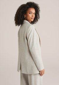 Helehall hall kohandatud bleiser õrna tekstuuriga, sälguga krae ja ühe tagumise lõhikuga, kantud üle särgi, lihtsate ehetemärgenditega.