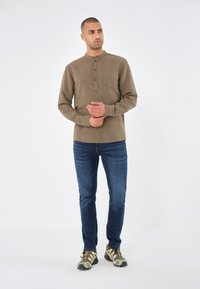 Braunes Langarmhemd mit Knöpfen am Hals, ausgestattet mit einer Brusttasche. Kombiniert mit dunkelblauen Jeans und Schuhen im Camouflage-Muster.