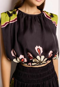 Blouse noire avec de grands motifs floraux en rose et vert. Présente un col et des manches froncés, avec des détails de taille élastique.
