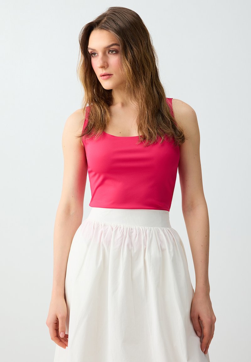 Jeune femme aux longs cheveux bruns portant un haut sans manches rose vif et une jupe blanche taille haute, regardant légèrement sur le côté.