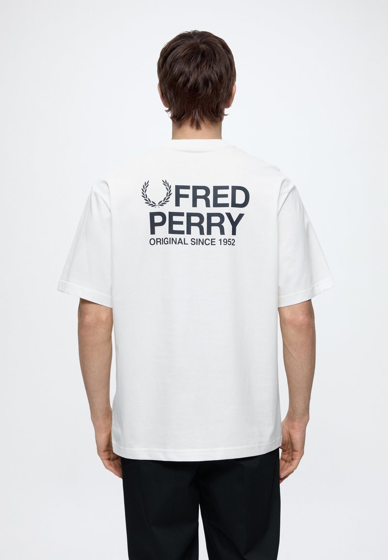 Άνδρας με λευκό T-shirt Fred Perry με μαύρο λογότυπο και κείμενο "Original since 1952" στην πλάτη, γυρνισμένος με την πλάτη προς το μέρος μας σε μονόχρωμο φόντο.