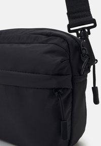 Czarna nylonowa torba crossbody z zapięciem na zamek, regulowanym paskiem, przednią kieszenią z sznurkiem do ciągnięcia oraz fakturowanym wykończeniem.