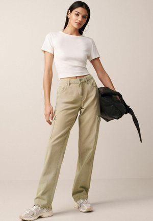 Světle zelené cargo kalhoty s bočními kapsami, doplněné o fitted bílé crop top. Modelka drží černou tašku a má na sobě bílé tenisky.