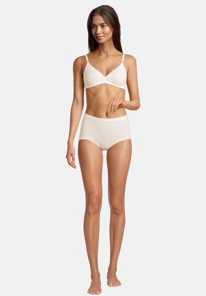 Woman standing barefoot wearing a beige wireless bra and high-waisted brief underwear against a plain white background.