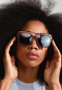 Ochelari de soare supradimensionați, de culoare marron, cu lentile întunecate cu gradient, un cadru transparent gros și accente pe brațe care contrastează cu părul texturat de culoare neagră.