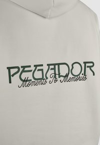 Biała bluza z kapturem z napisem "PEGADOR" dużymi zielonymi literami oraz "Moments To Memories" w czarnym kursywnym stylu na plecach.