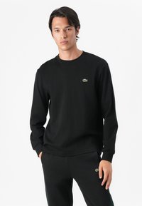 Black long-sleeve cotton t-shirt featuring a round neck and a small green Lacoste logo on the chest. Style is relaxed and fitted.