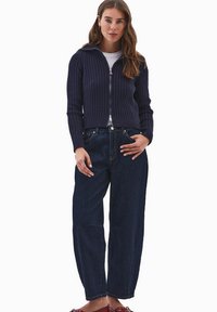 Tummansininen ribattu vetoketjulla varustettu huppari, jonka alla on valkoinen paita ja se on yhdistetty löysiin tummiin farkkuihin. Jalassa punaiset sandaalit.