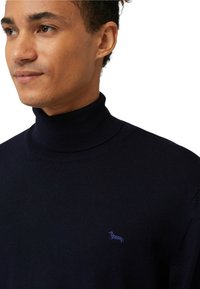Maglione a collo alto di colore blu navy in tessuto a maglia con un piccolo logo di un cane blu sul petto a sinistra. Il colletto è aderente per un aspetto avvolgente.