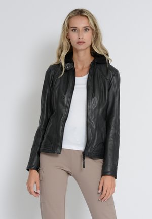 Black leather jacket features a zip-up front, hood, fitted design, and structured seams. Worn over a white top and beige pants.