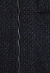 Dunkelblauer Wollmischstoff mit strukturierter Oberfläche, versehen mit einem zentralen schwarzen Reißverschluss und einem auffälligen Muster. Weiches und strukturiertes Design.