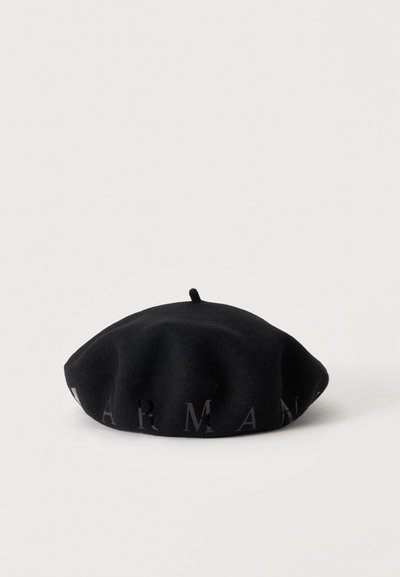 Béret en laine noire avec une petite tige sur le dessus et un lettrage sombre subtil autour de la base sur un fond blanc uni.